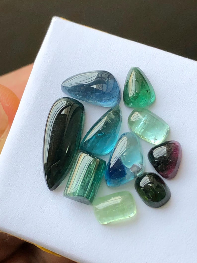 K&ouml;nnte beinhalten: Eine Sammlung polierter Edelsteine in verschiedenen Formen und Farben, darunter Blau, Gr&uuml;n und Schwarz. Die Edelsteine werden auf einer wei&szlig;en Oberfl&auml;che pr&auml;sentiert, die ihre durchscheinenden und reflektierenden Eigenschaften hervorhebt. Die Auswahl umfasst ovale, tropfenf&ouml;rmige und rechteckige Schnitte.