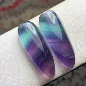 May include: Two elongated, polished fluorite gemstones. The stones display a gradient of colors, including purple, blue, and green. Each stone has a small hole at the top. The gemstones are displayed on a white surface.