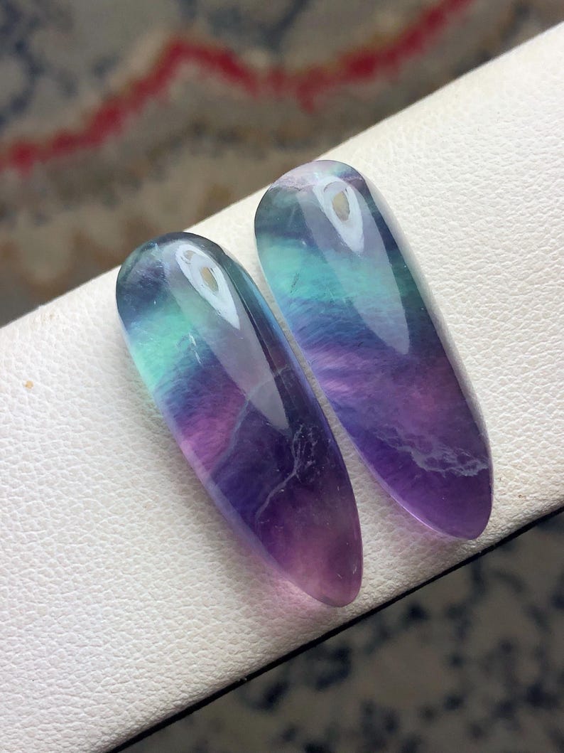 May include: Two elongated, polished fluorite gemstones. The stones display a gradient of colors, including purple, blue, and green. Each stone has a small hole at the top. The gemstones are displayed on a white surface.
