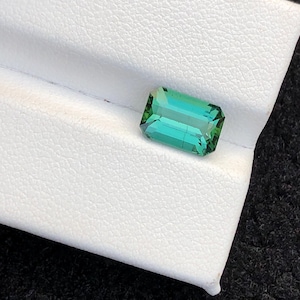 Puede incluir: Una gema rectangular con corte esmeralda, de un vibrante tono verde-turquesa. La gema se encuentra sobre una superficie blanca texturizada, mostrando sus facetas y claridad. El color de la gema transita de un verde intenso a un turquesa más claro.