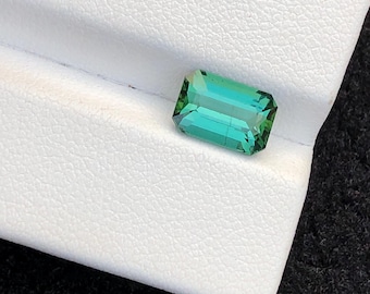 May include: A rectangular, emerald-cut gemstone with a vibrant teal-green hue. The gem is set against a white, textured surface, showcasing its facets and clarity. The gemstone's color transitions from a deep green to a lighter teal.
