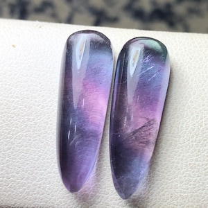 May include: Two elongated, polished fluorite stones. The stones display a gradient of colors, including purple, lavender, and hints of blue and green. The stones have a smooth, glossy surface and are set against a white background.