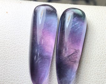 May include: Two elongated, polished fluorite stones. The stones display a gradient of colors, including purple, lavender, and hints of blue and green. The stones have a smooth, glossy surface and are set against a white background.