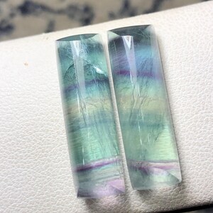 May include: Two rectangular fluorite gemstones. The stones are translucent with layers of green, blue, and purple hues. The gemstones have a polished, smooth surface and a rectangular shape with slightly angled edges.