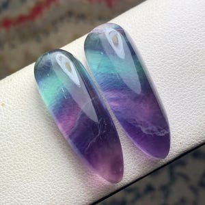May include: Two polished fluorite gemstones. The stones are elongated ovals, displaying a gradient of colors including purple, green, and blue. The gemstones are set against a white surface, with a blurred background.