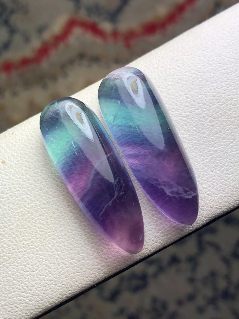 May include: Two polished fluorite gemstones. The stones are elongated ovals, displaying a gradient of colors including purple, green, and blue. The gemstones are set against a white surface, with a blurred background.