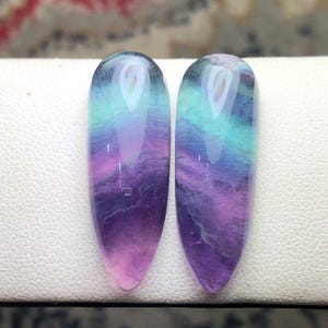 May include: Two polished, teardrop-shaped fluorite gemstones featuring a smooth, translucent surface with distinct color banding in shades of deep purple, vibrant magenta, and bright teal, arranged side by side in a vertical orientation.