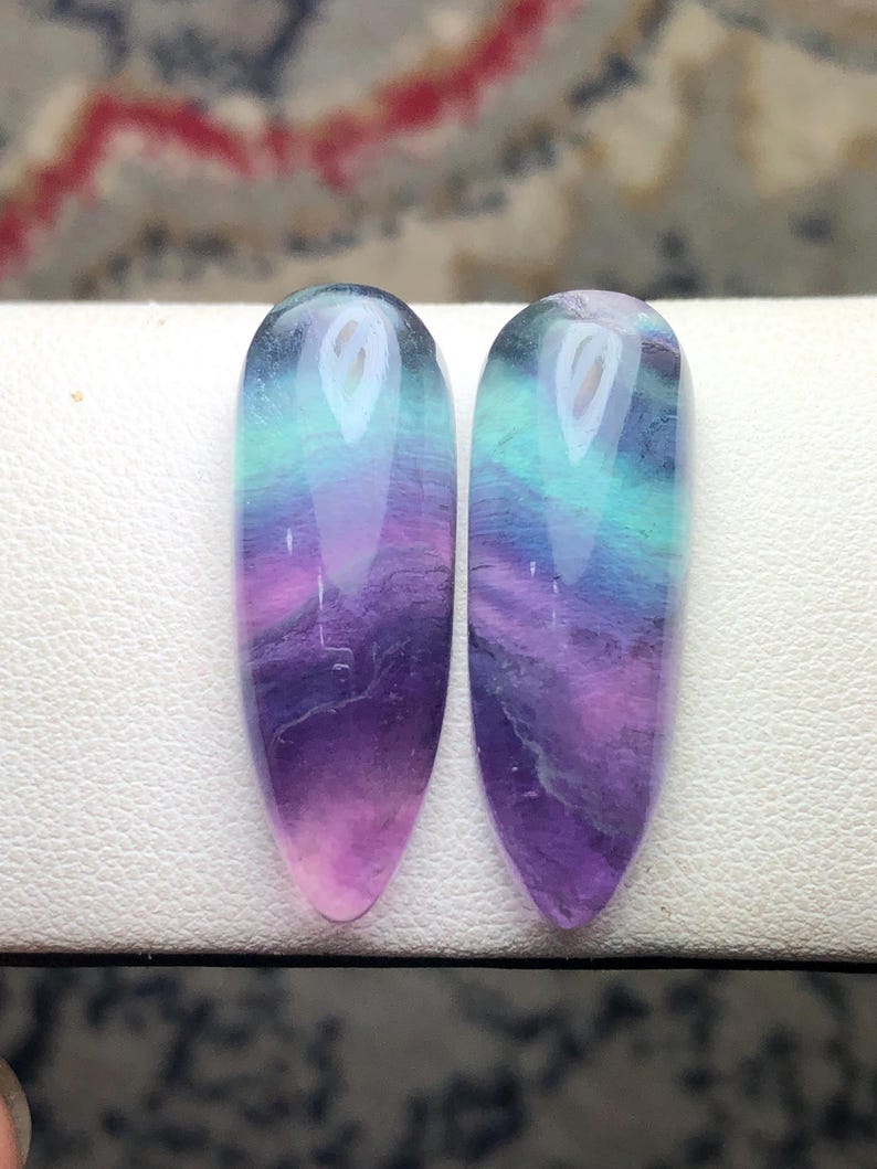 May include: Two polished, teardrop-shaped fluorite gemstones featuring a smooth, translucent surface with distinct color banding in shades of deep purple, vibrant magenta, and bright teal, arranged side by side in a vertical orientation.