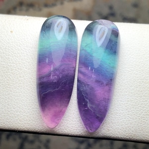 May include: Two polished, elongated gemstones with a gradient of colors. The stones transition from a light pink at the base, through shades of purple, to teal and blue at the top. The stones are smooth and reflective.