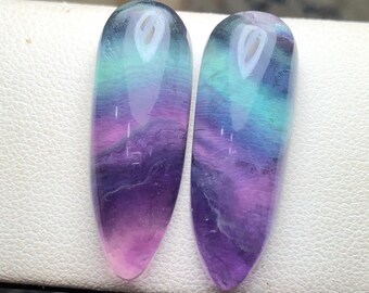 May include: Two polished, elongated gemstones displaying a gradient of colours. The stones transition from light pink at the base, through shades of purple, to teal and blue at the top. The stones are smooth and reflective.