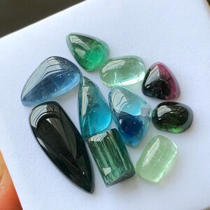 K&ouml;nnte beinhalten: Eine Sammlung polierter Edelsteine in verschiedenen Formen und Farben, darunter Blau, Gr&uuml;n und Schwarz. Die Edelsteine werden auf einer wei&szlig;en Oberfl&auml;che pr&auml;sentiert, die ihre durchscheinenden und reflektierenden Eigenschaften hervorhebt. Die Sammlung umfasst Turmalin und andere Edelsteine.