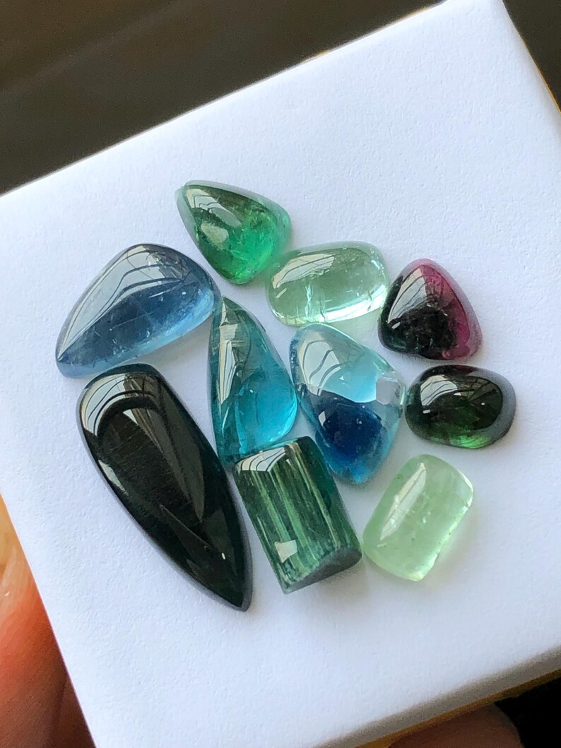K&ouml;nnte beinhalten: Eine Sammlung polierter Edelsteine in verschiedenen Formen und Farben, darunter Blau, Gr&uuml;n und Schwarz. Die Edelsteine werden auf einer wei&szlig;en Oberfl&auml;che pr&auml;sentiert, die ihre durchscheinenden und reflektierenden Eigenschaften hervorhebt. Die Sammlung umfasst Turmalin und andere Edelsteine.