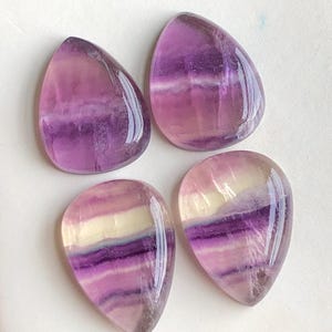 May include: Four polished teardrop-shaped fluorite gemstones. The stones display bands of purple, lavender, and cream colors. The translucent stones have a glossy finish and are arranged on a white surface.