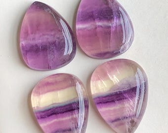 May include: Four polished, teardrop-shaped fluorite gemstones. The stones display bands of purple, lavender, and cream colours. The translucent stones have a glossy finish and are arranged on a white surface.