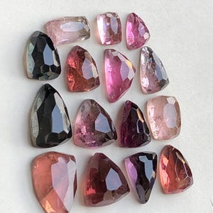 Puede incluir: Una colección de gemas facetadas en varias formas y tonos de rosa, morado y negro. Las gemas están dispuestas sobre una superficie blanca, mostrando sus superficies pulidas y claridad interna. El surtido incluye piedras rectangulares, triangulares y en forma de escudo.