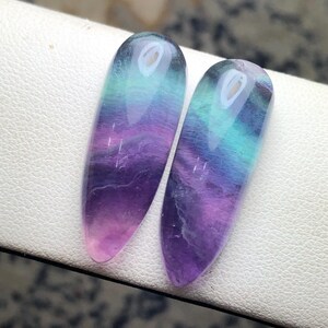 May include: Two polished fluorite stones, each with a gradient of colors. The stones transition from a translucent pink at the base to purple, then teal, and finally a clear top. The elongated shape is smooth and glossy.