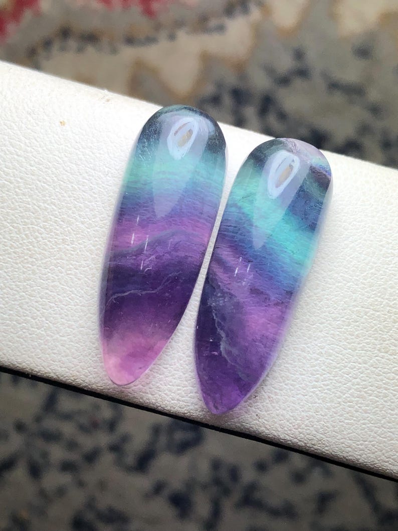 May include: Two polished fluorite stones, each with a gradient of colors. The stones transition from a translucent pink at the base to purple, then teal, and finally a clear top. The elongated shape is smooth and glossy.