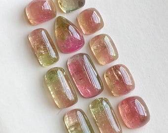 Pode incluir: Uma coleção de pedras preciosas de turmalina polidas em várias formas. As pedras exibem uma gama de cores, incluindo rosa, verde e amarelo, com uma qualidade translúcida. As pedras preciosas estão dispostas sobre uma superfície branca.