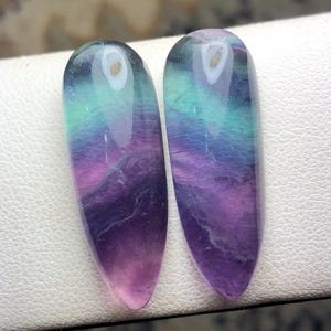 May include: Two polished fluorite gemstones. Each stone is a long, oval shape with a gradient of colors, including purple, pink, teal, and clear. The stones are displayed on a white surface. These gemstones are ideal for jewelry making or collecting.
