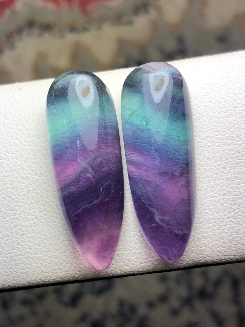 May include: Two polished fluorite gemstones. Each stone is a long, oval shape with a gradient of colors, including purple, pink, teal, and clear. The stones are displayed on a white surface. These gemstones are ideal for jewelry making or collecting.