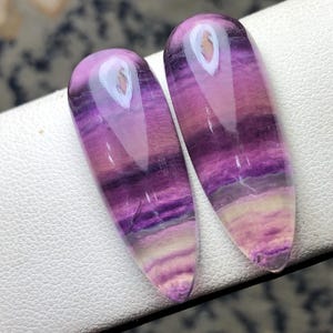 May include: Two polished, elongated gemstones with a gradient of purple, lavender, and white hues. The stones have a smooth, glossy surface and a teardrop shape, ideal for jewelry making or collecting. The gemstones are displayed on a white surface.