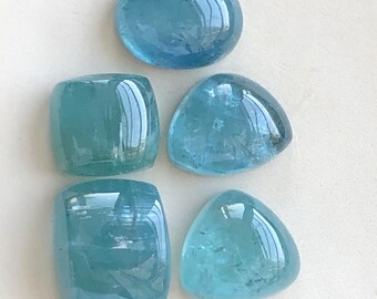 Pode incluir: Cinco pedras preciosas azul-claras, polidas e translúcidas, dispostas em grupo. A coleção inclui um cabochão oval, dois retangulares e dois triangulares, todos com superfícies lisas e arredondadas que refletem a luz com brilho.