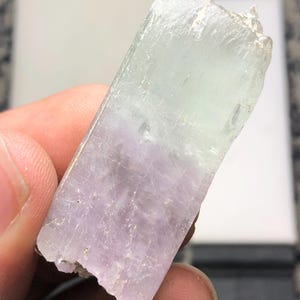 Pode incluir: Um cristal bruto, retangular, com um gradiente de cores. A metade superior é verde pálido, que transita para um lavanda suave na parte inferior. O cristal tem uma textura áspera e natural.