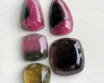 Pode incluir: Cinco pedras preciosas facetadas em tons de rosa, preto e amarelo-esverdeado. As pedras variam em forma, incluindo alongadas, quadradas e em forma de gota. As gemas são exibidas em uma superfície branca, mostrando suas superfícies polidas e cores internas.