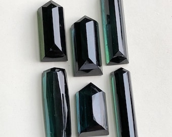 Pode incluir: Seis pedras preciosas polidas e facetadas em tons de verde-azulado escuro e preto. As pedras preciosas são retangulares e hexagonais, com pontas pontiagudas. As gemas estão dispostas sobre uma superfície clara, mostrando suas qualidades reflexivas.