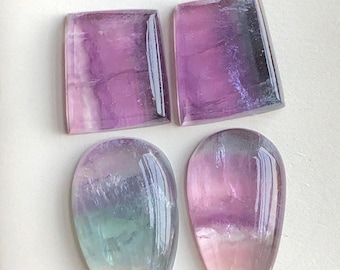 May include: Four polished fluorite gemstones in shades of purple, pink, and green. Two are rectangular, and two are teardrop-shaped. The stones have a translucent quality, with visible internal patterns and colour variations.