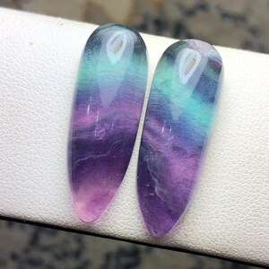 May include: Two polished, teardrop-shaped fluorite gemstones featuring a smooth, glossy finish. The stones display a vibrant gradient of colors, transitioning from deep purple at the pointed base to bright teal and light green at the rounded top.