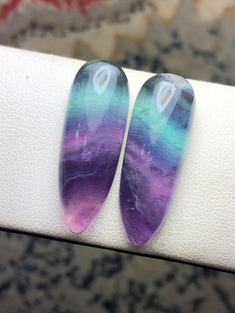 May include: Two polished, teardrop-shaped fluorite gemstones featuring a smooth, glossy finish. The stones display a vibrant gradient of colors, transitioning from deep purple at the pointed base to bright teal and light green at the rounded top.