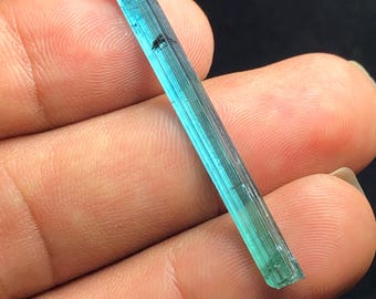 Può includere: Una gemma traslucida e allungata con un gradiente di tonalità blu e verdi. Il cristallo è tenuto in una mano, mostrando la sua forma naturale e grezza. La gemma ha una superficie leggermente ruvida e strutturata, con una piccola inclusione.