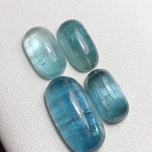 May include: Four oval-shaped, polished gemstones in varying shades of blue and teal. The stones have a smooth, glossy surface and are arranged on a white, textured background. The gems appear translucent, with internal striations.