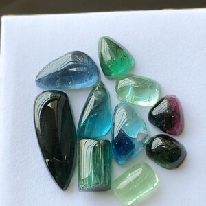 K&ouml;nnte beinhalten: Eine Sammlung polierter Edelsteine in verschiedenen Formen und Farben, darunter Blau, Gr&uuml;n und Schwarz. Die Edelsteine sind auf einer wei&szlig;en Oberfl&auml;che angeordnet, was ihre durchscheinenden und reflektierenden Eigenschaften hervorhebt. Die Sammlung umfasst Turmalin und andere Edelsteine.