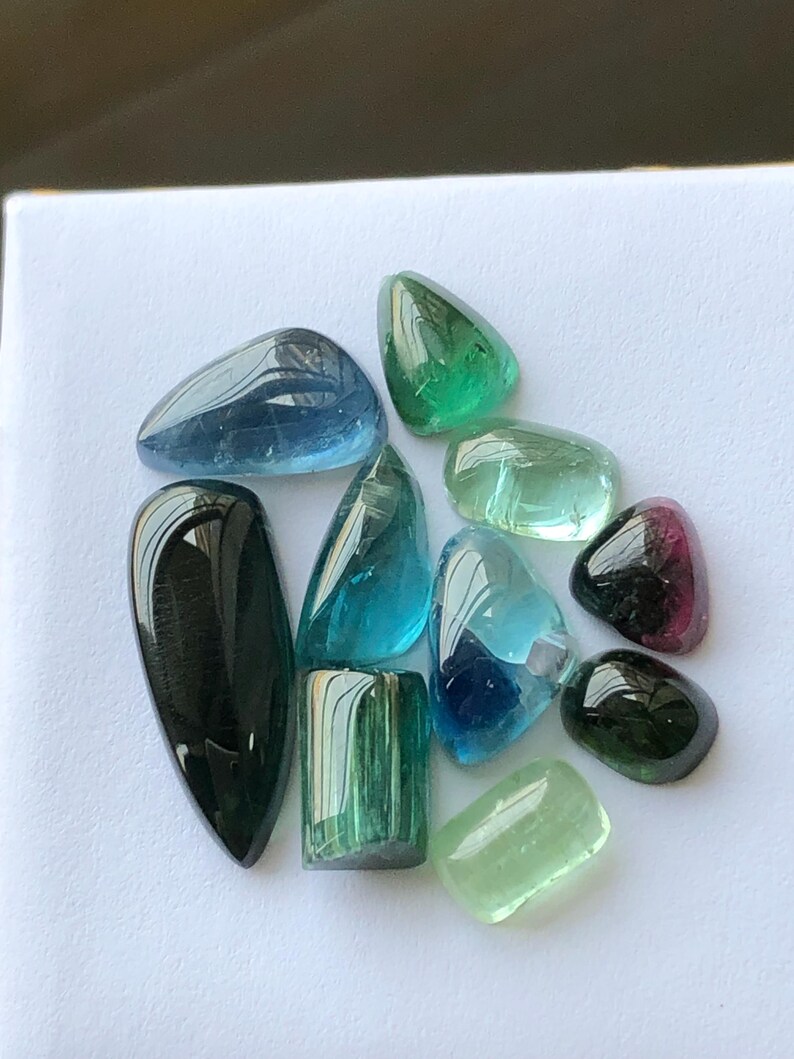 K&ouml;nnte beinhalten: Eine Sammlung polierter Edelsteine in verschiedenen Formen und Farben, darunter Blau, Gr&uuml;n und Schwarz. Die Edelsteine sind auf einer wei&szlig;en Oberfl&auml;che angeordnet, was ihre durchscheinenden und reflektierenden Eigenschaften hervorhebt. Die Sammlung umfasst Turmalin und andere Edelsteine.