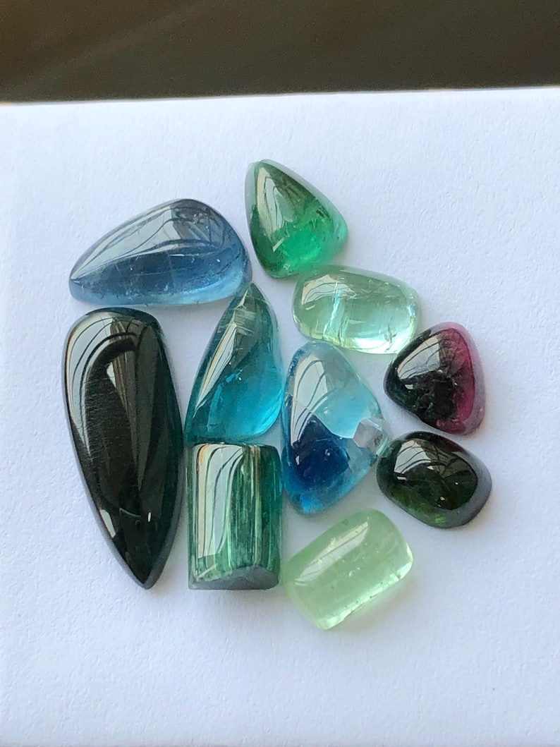 K&ouml;nnte beinhalten: Eine Sammlung polierter Edelsteine in verschiedenen Formen und Farben, darunter Blau, Gr&uuml;n und Schwarz. Die Edelsteine sind auf einer wei&szlig;en Oberfl&auml;che angeordnet, was ihre durchscheinenden Eigenschaften und ihre nat&uuml;rliche Sch&ouml;nheit hervorhebt. Die Sammlung umfasst Tropfen- und runde Formen.