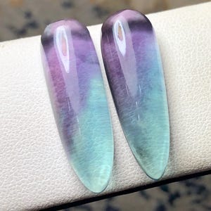 May include: Two polished, elongated gemstones with a gradient of colors. The stones transition from a deep purple at the top to a light blue-green at the bottom. The stones are smooth and translucent, with a glossy finish.