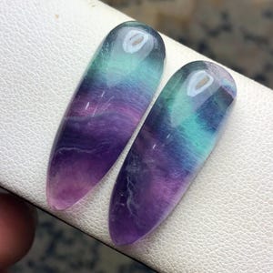 May include: Two polished, teardrop-shaped fluorite gemstones featuring a smooth, translucent surface with blended bands of deep purple, vibrant teal, and clear white. The stones exhibit a natural color gradient and a glossy finish.