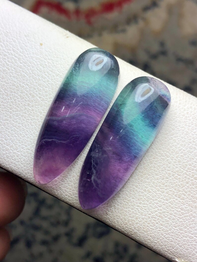 May include: Two polished, teardrop-shaped fluorite gemstones featuring a smooth, translucent surface with blended bands of deep purple, vibrant teal, and clear white. The stones exhibit a natural color gradient and a glossy finish.