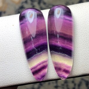 May include: Two polished, teardrop-shaped fluorite stones. The stones display bands of purple, pink, and cream colors. The translucent stones have a glossy finish and are set against a white surface.