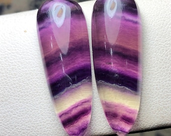 May include: Two polished, teardrop-shaped fluorite stones. The stones display bands of purple, pink, and cream colours. The translucent stones have a glossy finish and are set against a white surface.