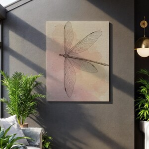 May include: A canvas art print featuring a detailed illustration of a dragonfly. The dragonfly is depicted in a brown line drawing against a soft, watercolor-style background with pink and yellow hues. The artwork is displayed on a dark gray wall.