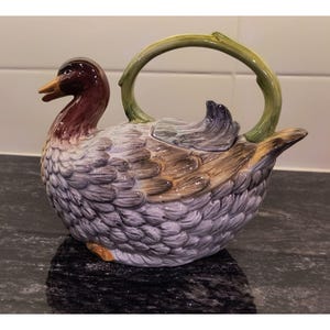 May include: A ceramic teapot shaped like a duck, featuring a detailed design. The duck's head is a deep burgundy, with a yellow beak. The body is a mix of gray, brown, and white, with a green handle. The teapot is designed to resemble a duck.