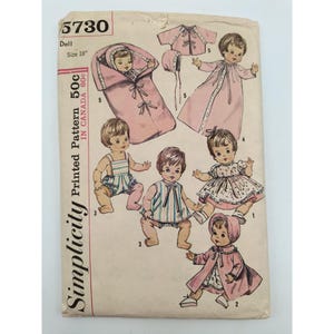 May include: A Simplicity sewing pattern envelope, number 5730, for an 18-inch doll. The front features illustrations of dolls wearing various outfits, including a pink bunting, a long pink gown, a dress, a jacket, and a striped romper with a bonnet.