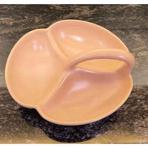 May include: A peach-colored, three-sectioned serving dish with a handle. The dish has a rounded, petal-like shape with a smooth, glossy finish. The handle is arched and integrated into the design, providing a comfortable grip.