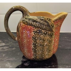 May include: A ceramic pitcher with a large handle and a spout. The pitcher is decorated with a floral pattern in shades of red, green, and brown. A leaf design is on the side of the pitcher.