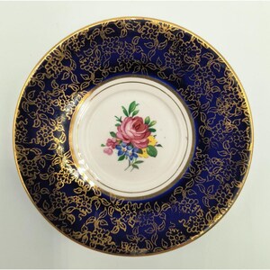 May include: A round ceramic saucer featuring a deep blue border decorated with a gold floral pattern. The center is white and displays a small, colorful bouquet of pink, yellow, and blue flowers. The rim is finished with a thin gold edge.