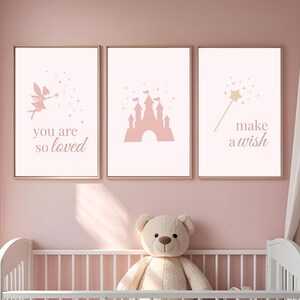 May include: Three framed prints with a fairy, castle, and wand design in pink and gold tones. The prints feature the phrases "you are so loved" and "make a wish". The prints are displayed above a white cot with a teddy bear.