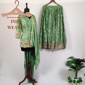 Può includere: Un completo abito etnico indiano verde, composto da top, pantaloni e gonna. Il top e la gonna sono impreziositi da dettagli dorati e rossi. L'abito è esposto su un manichino e su una gruccia.